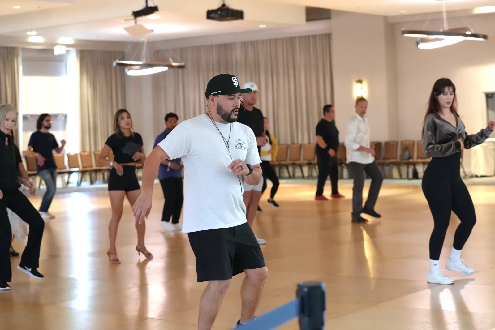 A group practises Bachata footwork in a bright dance hall.
