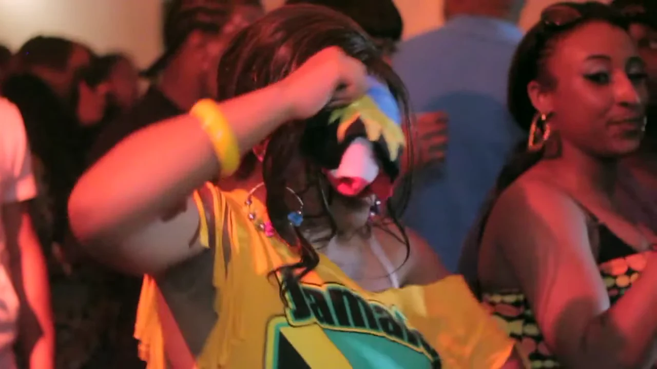 Londono dancehall vakarėlio dalyvė, apsirengusi geltonai, pakelia ryškių spalvų audinį prie veido.